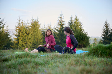 Two athletic young women dressed in sporty attire engage in a range of yoga poses amidst the scenic beauty of mountainous surroundings. The gentle rays of the morning sun cast on their slender bodies