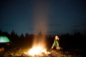 A young female traveler sits by the campfire near her tent, enjoying a moment of relaxation beneath...