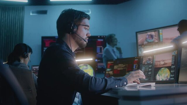 Flight control employee in headset sits in front of computers in command center, monitors space mission with astronaut. Team clap hands after space rocket launch displayed on big digital screens.