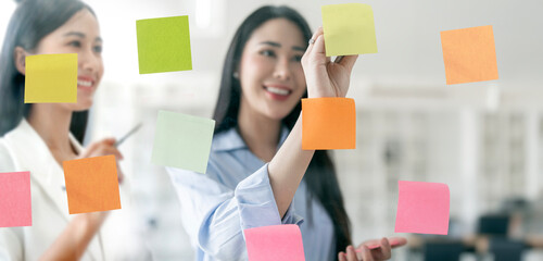 Young Creative businesswoman writing on sticky notes on a glass wall, female colleague looking.