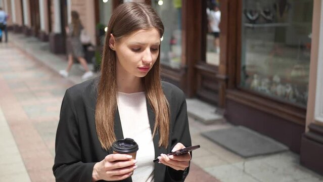 Beautiful young woman with takeaway drink using mobile phone while walking near building. Camera moving backwards, slow motion effect