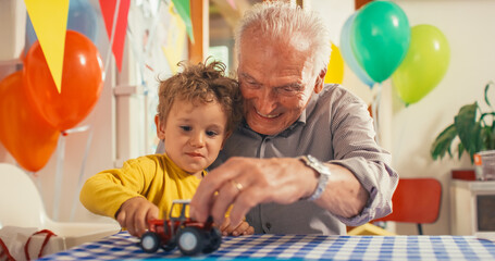 Fototapeta na wymiar Heartwarming Moment Shared Between a Senior Man and his Grandson: Happy Toddler and his Grandpa Playing with Truck and Plane Toys Together in a Kitchen Decorated with Balloons for a Birthday Party