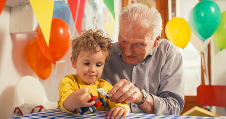 Fototapeta na wymiar Heartwarming Moment Shared Between a Senior Man and his Grandson: Happy Toddler and his Grandpa Playing with Truck and Plane Toys Together in a Kitchen Decorated with Balloons for a Birthday Party