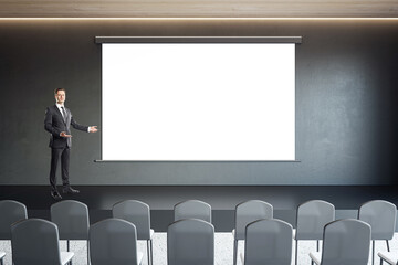 Business speaker on the scene near blank white screen for advertising text, campaign or logo brand on dark wall background in front of black chairs, marketing and lecture concept, mock up