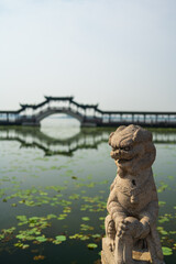 Fototapeta na wymiar Chinese traditional style stone lion with wooden bridge and lotus pool in background