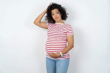 young pregnant woman wearing striped t-shirt over white background being confused and wonders about something. Holding hand on head, uncertain with doubt. Pensive concept.