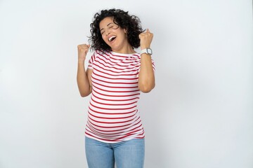 Ecstatic young pregnant woman wearing striped t-shirt over white background shout loud yeah fist up...