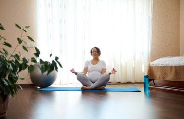 Authentic relaxed pregnant woman meditating in lotus pose with mudra gesture on yoga mat, in cozy home bedroom, against large window on sunny day. Healthy active lifestyle, wellness in pregnancy time - obrazy, fototapety, plakaty