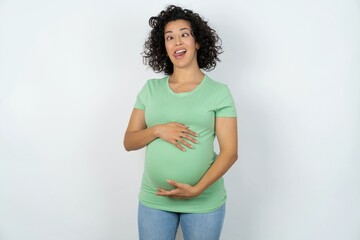 Funny young pregnant woman wearing green t-shirt over white background makes grimace and crosses...