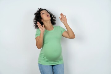 young pregnant woman wearing green t-shirt over white background keeps palms forward and looks with...