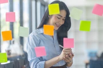 Smiling business Asian people thinking and use sticky notes on glass wall in office with her colleague in background.
