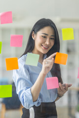 Smiling business Asian people thinking and use sticky notes on glass wall in office with her...