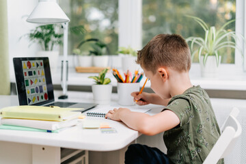 Distance learning online education. Caucasian smile kid boy studying at home with laptop and doing school homework. Thinking child siting with notebook, pencils and training books. Back to school.