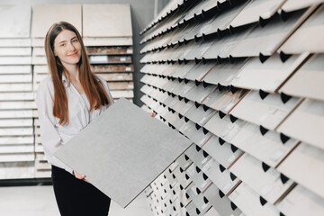 Seller Consultant young woman shows sample of gray ceramic tiles for loft style, refurbishment in...