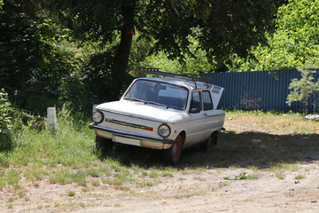A white retro subcompact car, Soviet-made in the 1970s, parked on the side of a sandy road near a lake.
