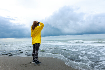Nervous young woman in yellow raincoat standing alone by the sea on a rainy day