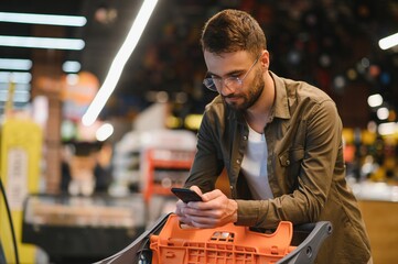 Quick text during shopping. Handsome young man holding mobile phone and smiling while standing in a food store