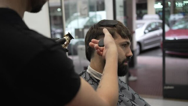 Close-up video of a barber cutting the sideburns and doing a gradient to his client with a hair clipper and cleaning him with a small brush.