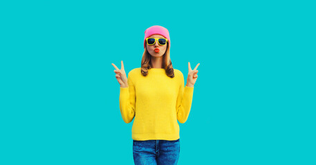 Stylish modern young woman model 20s blowing her lips sending sweet air kiss wearing colorful clothes, yellow knitted sweater and pink hat on blue background