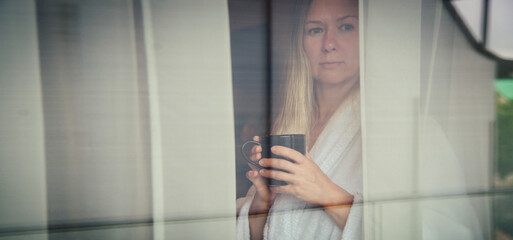 Fototapeta na wymiar Woman in elegant robe drinking coffee in hotel room and standing near window. window reflection