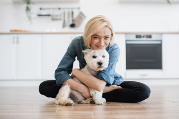 Portrait of smiling woman hugging gently adorable dog while posing on wooden floor with legs crossed. Happy female owner showing care for beloved pet before going for after dinner walk to park.