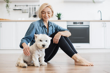 Portrait of cheerful young lady sitting next to well-behaved white dog on bare floor in kitchen...