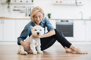 Joyful young woman in yoga pants rubbing pet coat while seating on room flooring in spacious apartment. Playful talkative dog owner cheering small terrier on during home treasure hunt game indoors.
