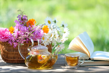 glass teapot and cup with herbal tea, flowers in basket, book close up on table, natural abstract...
