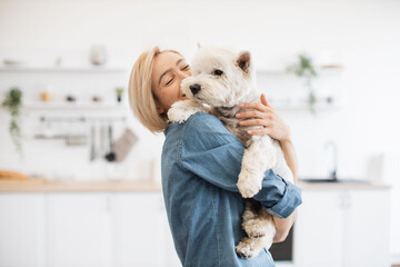 Beautiful woman in jeans shirt holding medium-sized dog on shoulder while standing in light room...