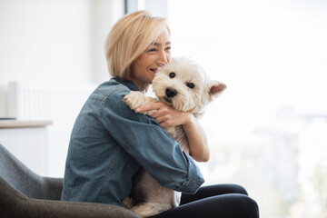 Close up view of cheerful caucasian lady in denim shirt snuggling into soft dog's coat while resting at home. Smiling young pet lover being overwhelmed with tenderness and care for small terrier.