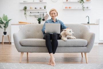 Full length view of barefoot woman looking at computer screen while touching white terrier on top...