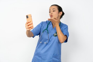 Young doctor woman wearing blue uniform over isolated background blows air kiss at camera of smartphone and takes selfie, sends mwah via online call.