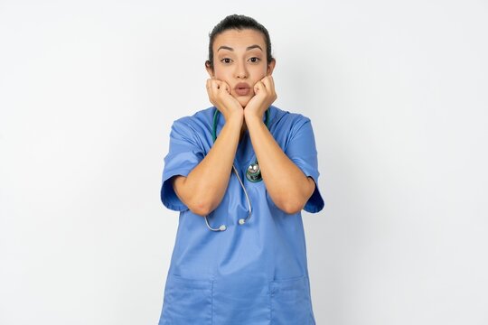 Young doctor woman wearing blue uniform over isolated background with surprised expression keeps hands under chin keeps lips folded makes funny grimace