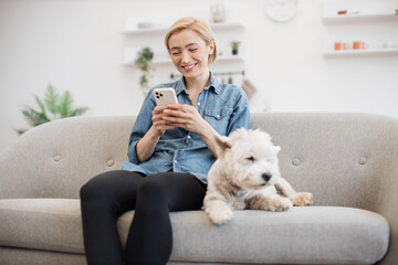 Attractive young woman in cozy outfit holding smartphone while her small pet having rest by her side on gray sofa. Active pet owner sharing online video of great outdoor fun with furry friend.