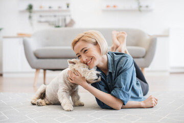 Pretty smiling woman in casual wear snuggling to cute pet's head while testing comfort of wooden...