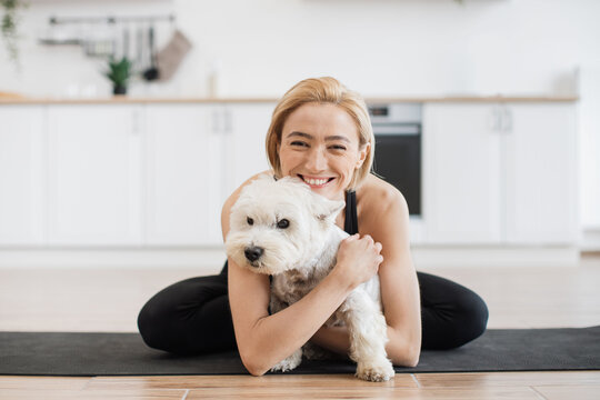 Affectionate adult female in yoga activewear embracing white terrier while sitting cross-legged on rubber mat indoors. Healthy lady awarding sweet pet with hug for good behaviour during home workout.