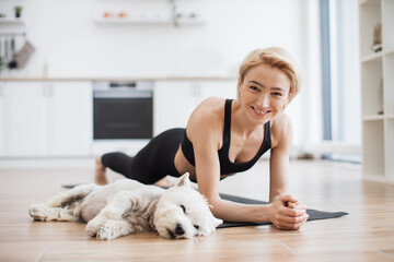 Peaceful blonde lady in black activewear doing yoga practice while tired Westie resting beside on floor of kitchen. Athletic yogini holding Forearm Plank Pose during yoga session with pet at home.