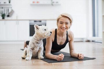 Portrait of happy blonde woman snuggling to adorable little dog while holding Forearm Plank Pose at...