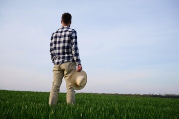 Rear view. Standing and holwing straw hat in hand. Young man is on agricultural field - obrazy, fototapety, plakaty