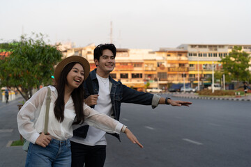 Happy smiling Young Asian couple traveler tourists hailing for taxi cab on the street in Bangkok in Thailand with no phone or use of technology