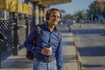 Young latin man with headphones waiting for the bus with a paper cup of coffee in hand.