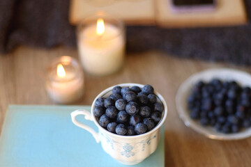 Cup of blueberries, set of books, phone, eyeglasses, lit candles and blanket on the table. Hygge at home. Selective focus.
