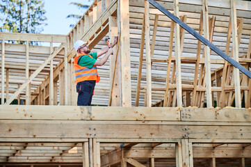 Carpenter constructing two-story wooden frame house near the forest. Bearded man hammering nails into structure while wearing protective helmet and construction vest. Concept of modern construction.