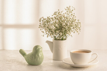 Coffee, a bouquet of gypsophila and a porcelain figurine of a bird on a white table. Women's desk. Cozy breakfast.