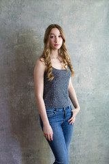 Beautiful slim young woman with long wavy blond hair while standing in tank top and jeans against concrete wall
