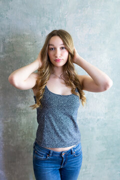Beautiful slim young woman touching her long wavy blond hair while standing in tank top and jeans against concrete wall