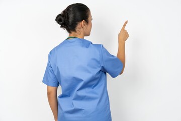 Beautiful doctor woman standing over white studio background pointing to object on copy space, rear view. Turn your back