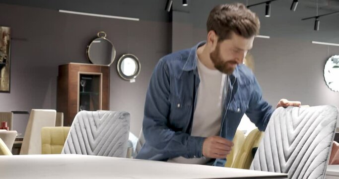 Side view of male consumer buying furniture, choosing chair in shooping mall, hugging, smiling. Handsome man wearing jeans shirt finding grey chair. Concept of urban lifestyle.
