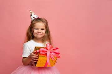little girl in party hat holds gift box in front of pink background