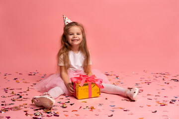 little girl in festive hat with gift box is sitting on floor on pink background